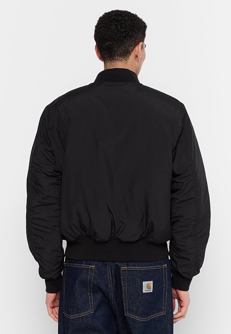 Personne de dos portant une veste bomber noire et un jean bleu foncé avec un patch logo Carhartt sur la poche arrière.