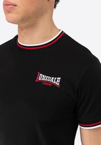 Must mustre emane ümmarguse kaelusega, millel on punase ja valge triibuga aktsendid. Rinnal on tikandiga "Lonsdale London" logo.