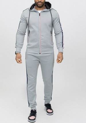 Survêtement gris avec une veste à zip et un pantalon assorti. Présente des rayures rouges et bleu marine le long des manches et des côtés, fabriqué en tissu lisse.