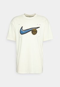 T-shirt bawełniany w kolorze kremowym z czarnym logo Nike oraz okrągłym logo Paryża w złotym i niebieskim kolorze, z małymi kolorowymi elementami dekoracyjnymi.
