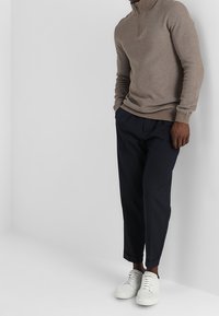 Beige Zip-Pullover, dunkelblaue Hose und weiße Sneakers. Der Pullover hat einen hohen Kragen und eine strukturierte Strickart. Die Hose hat eine maßgeschneiderte Passform.