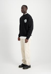 Felpa nera con dettagli grafici, indossata sopra una camicia bianca. Abbinata a pantaloni cargo beige e scarpe nere con cuciture gialle.