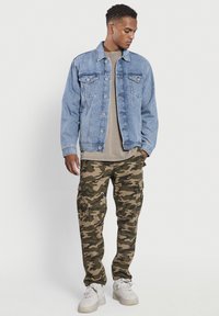 Lichtblauwe spijkerjacket met lange mouwen, knoopsluiting en twee borstzakken. Tan T-shirt eronder, camo cargobroek en witte sneakers.