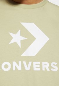 T-shirt en coton vert clair arborant un grand design d'étoile blanche et de flèche, avec le mot "CONVERSE" imprimé en blanc juste en dessous.