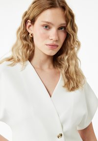 Weiße V-Ausschnitt-Bluse mit kurzen Ärmeln, die an der Vorderseite mit goldenen Knöpfen versehen ist. Die Haare sind in sanften, lockeren Wellen gestylt. Klare, blaue Augen.