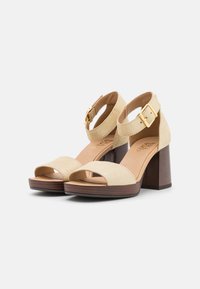 Beige Wildledersandalen mit offenem Zehenbereich und Knöchelriemen, mit einer goldenen Schnalle und einem Holzblockabsatz. Glatte Textur und schlichtes Design.