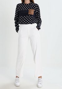 Maglietta a maniche lunghe navy con pois bianchi e taschino in pelle marrone, abbinata a pantaloni bianchi a gamba larga e sneakers bianche.