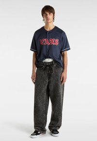 Marineblå baseballtrøje med rød "VANS" tekst, kombineret med løse, slidte sorte denimbukser og sorte sneakers med hvide detaljer.