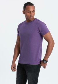 Lila kortärmad t-shirt i mjukt tyg med rund halsringning och avslappnad passform, bärs med svarta jeans och en klocka.