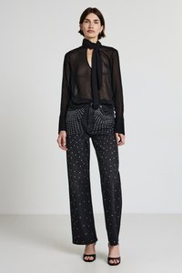 Schwarze transparente Bluse mit langen Ärmeln und einer Schleife am Hals, kombiniert mit hoch taillierten schwarzen Jeans, die mit silbernen metallischen Nieten in einem gepunkteten Muster verziert sind.