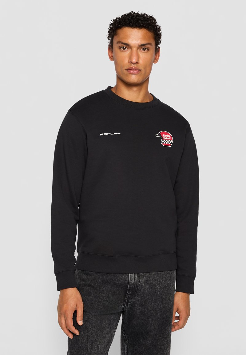 Man wearing black sweatshirt with "Replay" text and red helmet logo on chest, paired with dark gray jeans, standing against plain background.