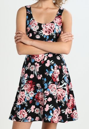 Femme portant une robe noire sans manches avec un motif floral rose, rouge et bleu, les bras croisés sur la poitrine devant un fond uni.