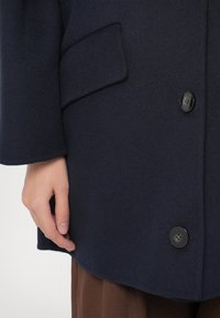 Sügavsinine villane mantel, millel on ümar alläär, taskud ja mustad nupud. Käsikäe nähtav asetus riide peal lisab tekstuurset detaile.