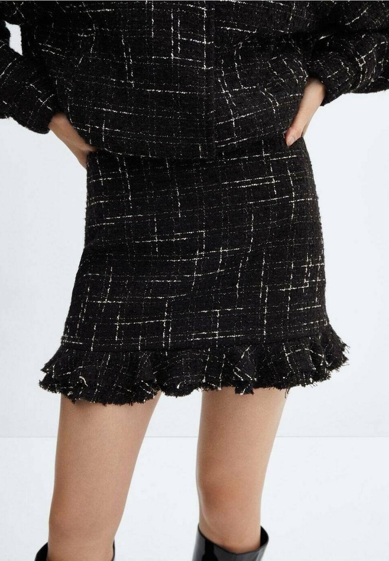 Mini-jupe en tweed noir à motif à carreaux blancs et ourlet volanté portée avec une veste assortie, mains du mannequin sur les hanches.