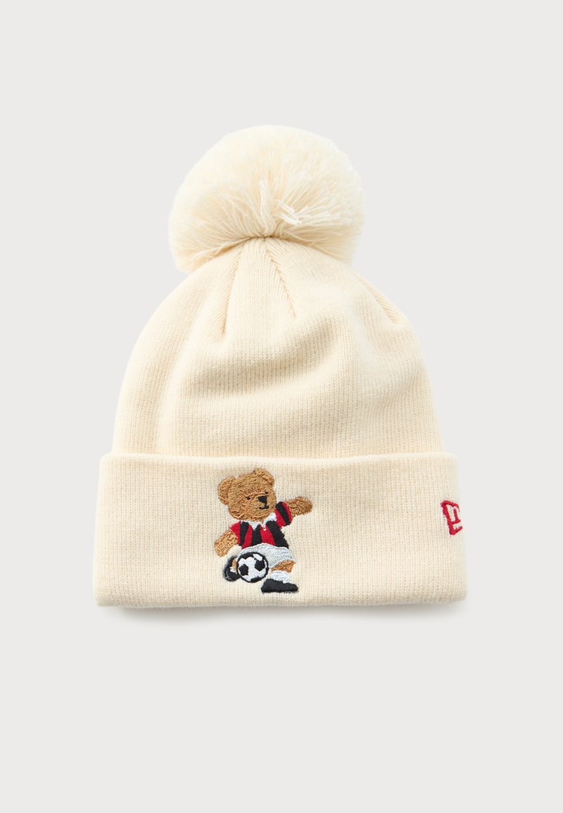 Bonnet en tricot crème avec pompon, présentant un ours en peluche brodé en train de frapper un ballon de football et un logo de marque rouge sur le revers.