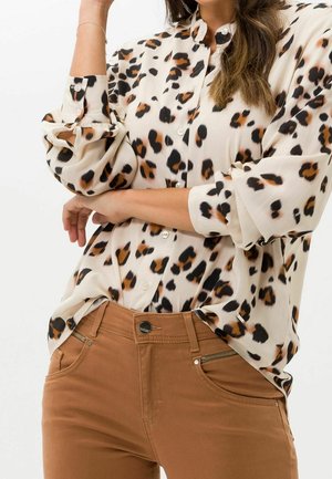 Kobieta ubrana w beżową bluzkę w panterkę z podwiniętymi rękawami oraz brązowe, wysokowalne spodnie z detalami na zamkach.