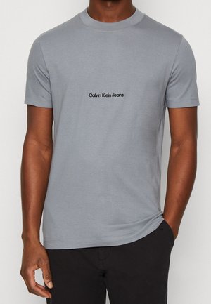 Mann trägt ein hellgraues T-Shirt mit kurzen Ärmeln und dem Schriftzug "Calvin Klein Jeans" zentriert auf der Brust, kombiniert mit schwarzen Hosen.