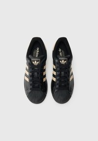 Pár černých tenisek Adidas s béžovými pruhy a špičkami ve tvaru ulity, zavázané, pohled zhora na jednobarevném bílém pozadí.