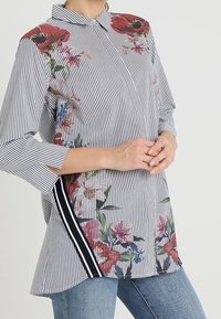 Koszula w paski z kwiatowym nadrukiem po jednej stronie, z kołnierzykiem, rękawami do łokcia i akcentem w postaci paska po boku. Materiał z mieszanki bawełny.