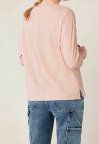 Helles rosa Langarmoberteil mit lässigem Schnitt, weichem Material, seitlichen Schlitzen und rundem Ausschnitt, kombiniert mit blauen Cargo-Hosen.