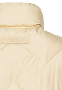Lys beige quiltet jakke med højt krave, synlige sømme og et mærke med brandnavnet tydeligt vist.