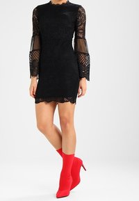 Robe en dentelle noire avec de longues manches transparentes en forme de cloche, ourlet festonné, associée à des talons de style chaussette rouges. Motif floral texturé sur l'ensemble.