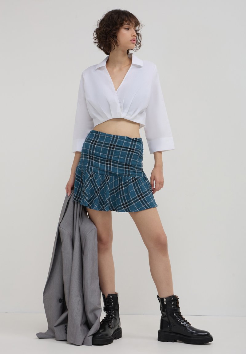 Camicia bianca tagliata, gonna corta a quadri color teal con balza, blazer grigio drappeggiato, anfibi neri stringati. Design semplice e linee pulite.
