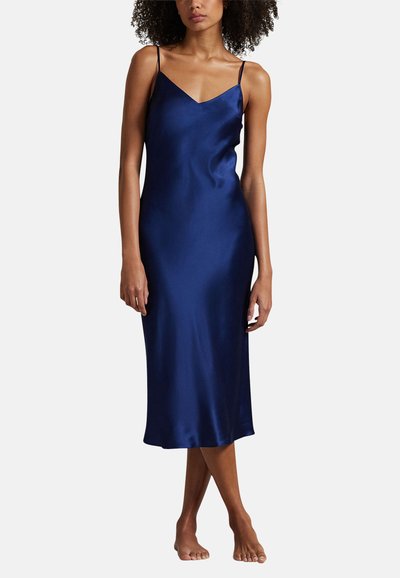 Vrouw die een mouwloze midi slipjurk van marineblauwe satijn draagt met dunne bandjes, blootsvoets staand tegen een effen achtergrond.