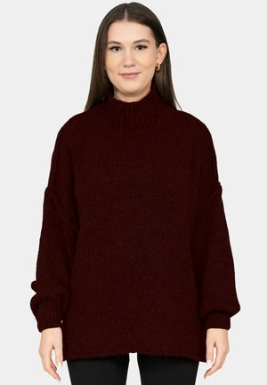 Junge Frau mit glatten braunen Haaren, die goldene Creolen und einen dunkelburgunderfarbenen Rollkragenpullover trägt, steht vor einem schlichten Hintergrund.