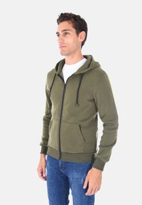 Felpa con zip di un verde oliva, dotata di tasca frontale, cappuccio regolabile con cordino e polsini a coste. Realizzata in un morbido tessuto texture.