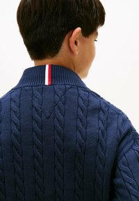 Tumši zilā adītā džemperī ar izcilu šņorējuma rakstu, ar ribotu apkakli un sarkano un baltās svītras akcentu aizmugurē.