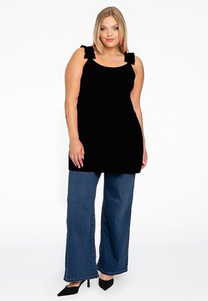 Schwarze Tunika mit Schleifenakzenten an den Schultern, kombiniert mit weiten blauen Jeans und schwarzen High Heels. Der Stoff ist glatt und der Schnitt entspannt.