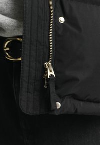 Zwarte gewatteerde jas met een gouden rits, voorzien van een drukknoopsluiting en textielstof. Zichtbare stiksels langs de randen en een logotab.