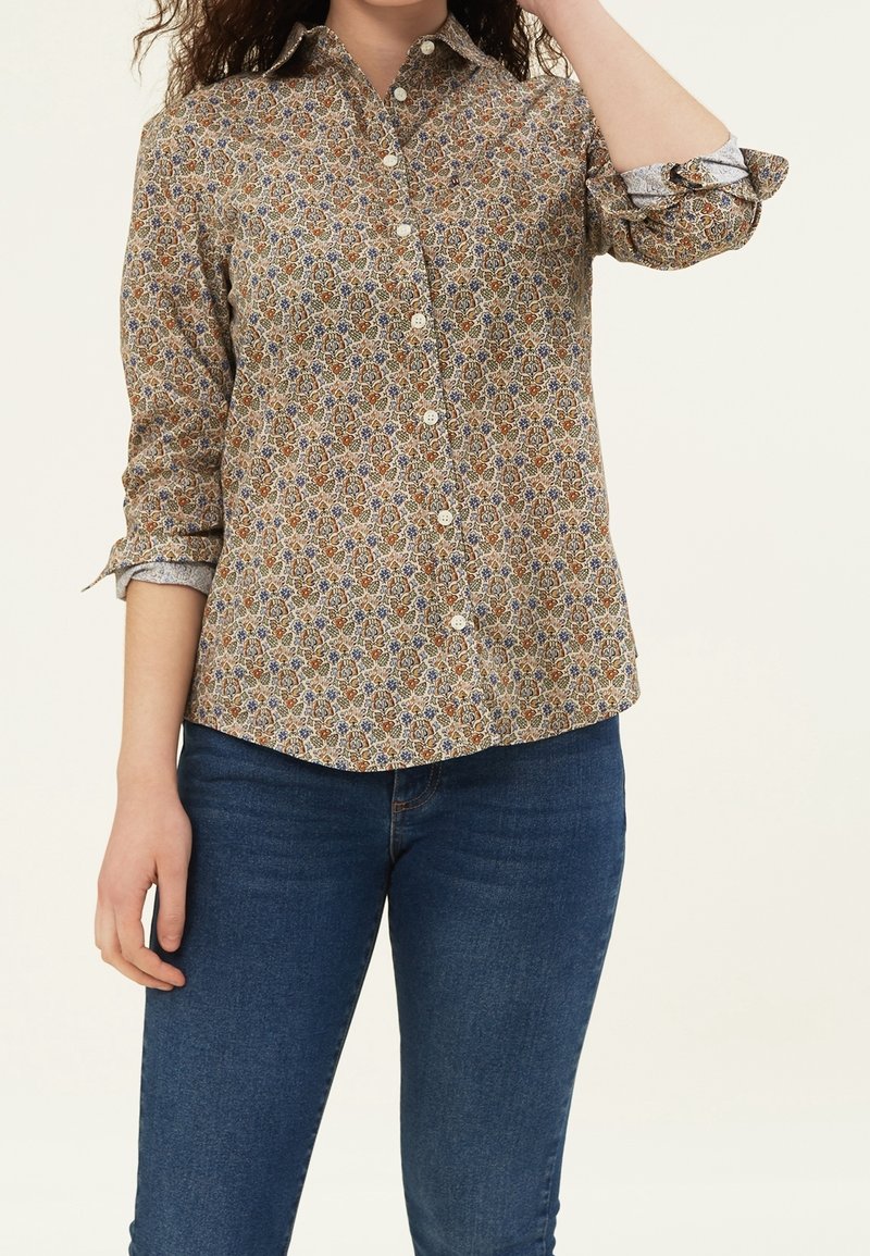 Femme portant une chemise beige à fleurs boutonnée avec manches retroussées et un jean bleu foncé sur un fond uni.