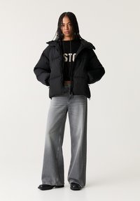 Giubbotto puffer nero con cappuccio, caratterizzato da un design trapuntato e due tasche frontali. Indossato sopra un top a maglia nera e abbinato a jeans grigi a gamba larga.