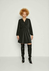 Γυναίκα με σγουρά μαλλιά που φοράει ένα μακρυμάνικο μαύρο φόρεμα με λαμπερή υφή και μαύρες μπότες με τακούνι μέχρι το γόνατο, στέκεται μπροστά σε έναν απλό ανοιχτό φόντο.