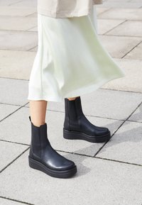 Botines de cuero negro hasta el tobillo con un acabado suave, paneles laterales elásticos y una suela de plataforma gruesa. Llevados con una falda ligera de color claro.