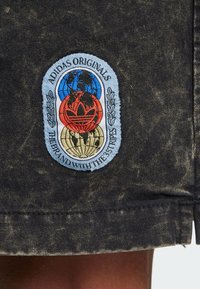 Svart denimtyg med en cirkulär Adidas Originals-logotyp som har blå, röda och gyllene element, omgiven av lagerblad och text.