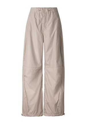 Jong persoon staand met armen over elkaar, draagt een zonnebril, een muts, een taupekleurig T-shirt, losse lichtbeige broek en beige sneakers op een witte achtergrond.