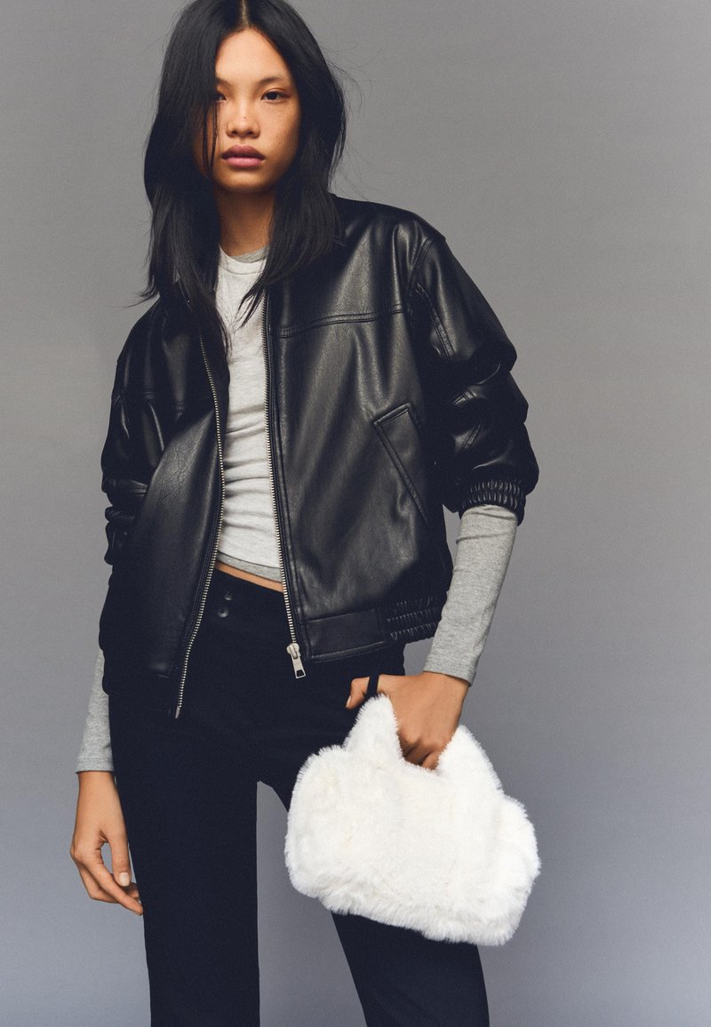 Crna kožna bomber jakna sa rajsferšlusom sprijeda, rebrastim manžetama, uparena sa svijetlo sivim topom i crnim farmerkama. Drži bijelu pahuljastu torbicu.