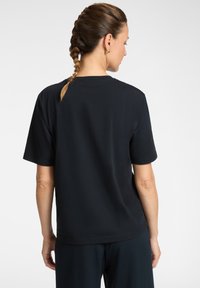 Femme aux cheveux tressés portant un t-shirt noir à manches courtes simple et un pantalon noir, vue de dos sur fond blanc.