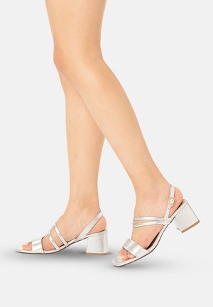 Kvinde ben iført sølvfarvede, åbne sko med blokhæl, tynde stropper og ankelrem med spænde på en lys baggrund.