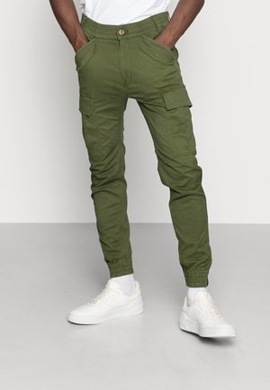 Groene cargobroek met tapse pijpen, elastische tailleband en meerdere zakken, gecombineerd met witte sneakers en zichtbare enkelmanchetten.