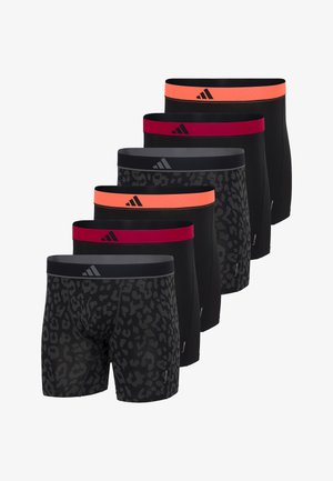 Athletischer Mann steht barfuß und trägt schwarze Boxer-Shorts mit orangenem Bund, posiert vor einem einfarbigen hellen Hintergrund.