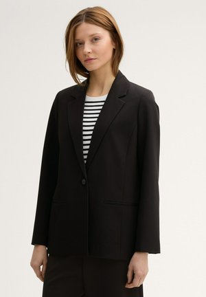 Schwarzes Blazer mit Reverskragen, Ein-Knopf-Verschluss, zwei Vordertaschen und langen Ärmeln, getragen über einem gestreiften Hemd.