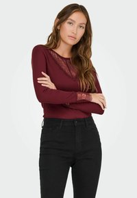 Bordeaux lange mouwen top met een kantenkraag en kantaccenten op de mouwen, gecombineerd met zwarte high-waisted jeans, beide aansluitend.