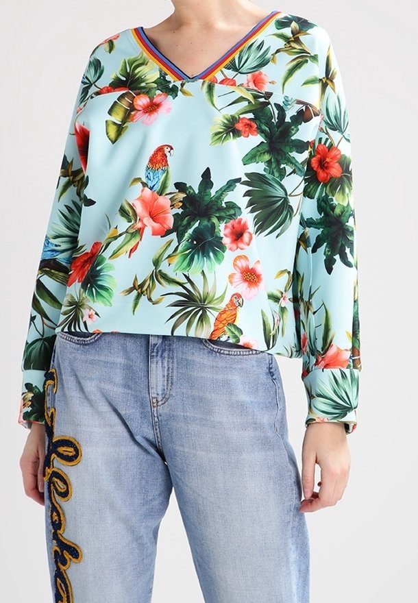 Světlemodrý top s dlouhým rukávem a květinovým vzorem, s výstřihem do V, zdobený jasnými papoušky a listy; v kombinaci s světlemodrými džíny s vyšíváním.