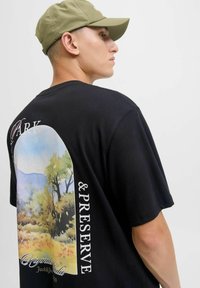 Vīrietis ar olīvu zaļu cepuri un melnu T-kreklu, kurā ir ainavains dabas attēls un teksts "Parks un Aizsardzība" aizmugurē.