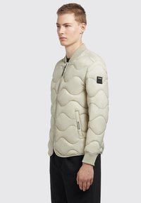 Ljust beige kviltad bomberjacka med v�ågmönster, ribbade muddarmar, sidofickor och dragkedja. Har en logopatch på ärmen.
