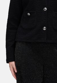 Czarna teksturowana kurtka z srebrnymi guzikami i kieszeniami, zestawiona z czarnymi błyszczącymi spodniami, pokazana od talii do połowy uda, lewa ręka spoczywa przy boku.